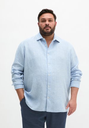 Man with beard wearing light blue long-sleeve button-up shirt and dark pants, standing with one hand in pocket against white background.