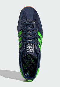 Adidași din suede marin cu dungi verzi, legare frontală cu șireturi și un logo pe o branț negru cu model. Design cu talpă din cauciuc rotunjită.