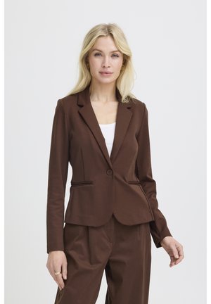 Heledate juustega naine, kes kannab kohandatud pruuni bleiserit ja sobivaid pükse valge topi kohal, seisab ühevärvilise heleda tausta ees.