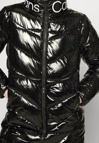 Svart puffjacka med en glänsande, kviltad yta. Har en hög krage, dragkedja och en kontrasterande vit logotyp vid nacken.