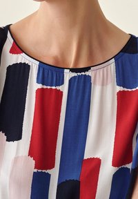 Gros plan sur le cou et le haut de la poitrine d'une femme portant un t-shirt sans manches avec un motif abstrait de rectangles rouges, bleus, noirs et blancs.