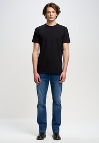 Camiseta de algodón negra con cuello redondo, combinada con jeans rectos azules. El modelo se encuentra en un entorno de estudio minimalista.