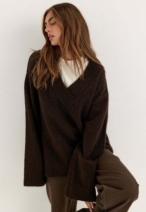 Giovane donna con lunghi capelli castani che indossa un maglione a V marrone scuro oversize sopra una camicia bianca e pantaloni marroni, che posa davanti a uno sfondo bianco.