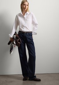Camisa blanca de botones con mangas largas, combinada con unos vaqueros de pierna ancha en azul oscuro. Lleva un bolso marrón y una bufanda estampada.