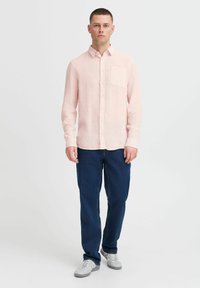 Chemise boutonnée rose clair avec une poche poitrine, associée à un jean bleu foncé et des baskets grises. Texture lisse, coupe décontractée.