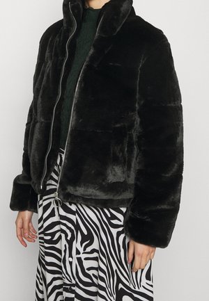 Personne portant une veste zippée en fausse fourrure noire par-dessus un haut vert foncé et une jupe imprimée zèbre noir et blanc.