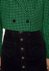 Zeleno-černé sako s vzorem psího zubu a dvojicí knoflíků, doplněné černou manšestrovou sukní s předními kapsami a zlatými kovovými prvky.