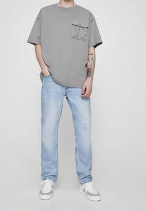 Personne portant un jean bleu clair, un T-shirt gris surdimensionné avec une poche poitrine, des baskets blanches et grises, dévoilant un tatouage coloré sur l'avant-bras gauche.