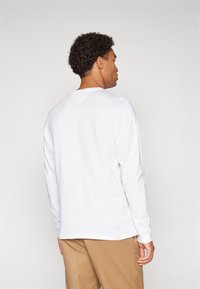 Camisa branca de manga longa com decote redondo e um detalhe em V subtil nas costas, combinada com calças castanhas claras.