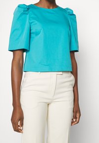 Femme portant un haut turquoise à manches bouffantes et un pantalon crème taille haute, debout devant un fond blanc uni.