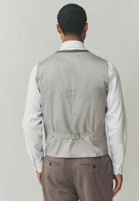 Graue Satinweste mit einem eleganten Design und verstellbarem Schnallen-Detail, getragen über einem weißen Hemd, ergänzt durch braune Hosen.