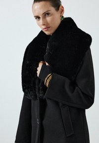 Vrouw in een zwarte jas met een grote zwarte nepbontkraag, gouden armbanden, een gouden ring en groen-gouden oorbellen tegen een effen achtergrond.