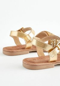 Goldene, metallische Sandalen mit flacher Sohle, die über kreuzweise Riemen, einen Schnallenverschluss und eine samtige Fußbett für zusätzlichen Komfort verfügen.