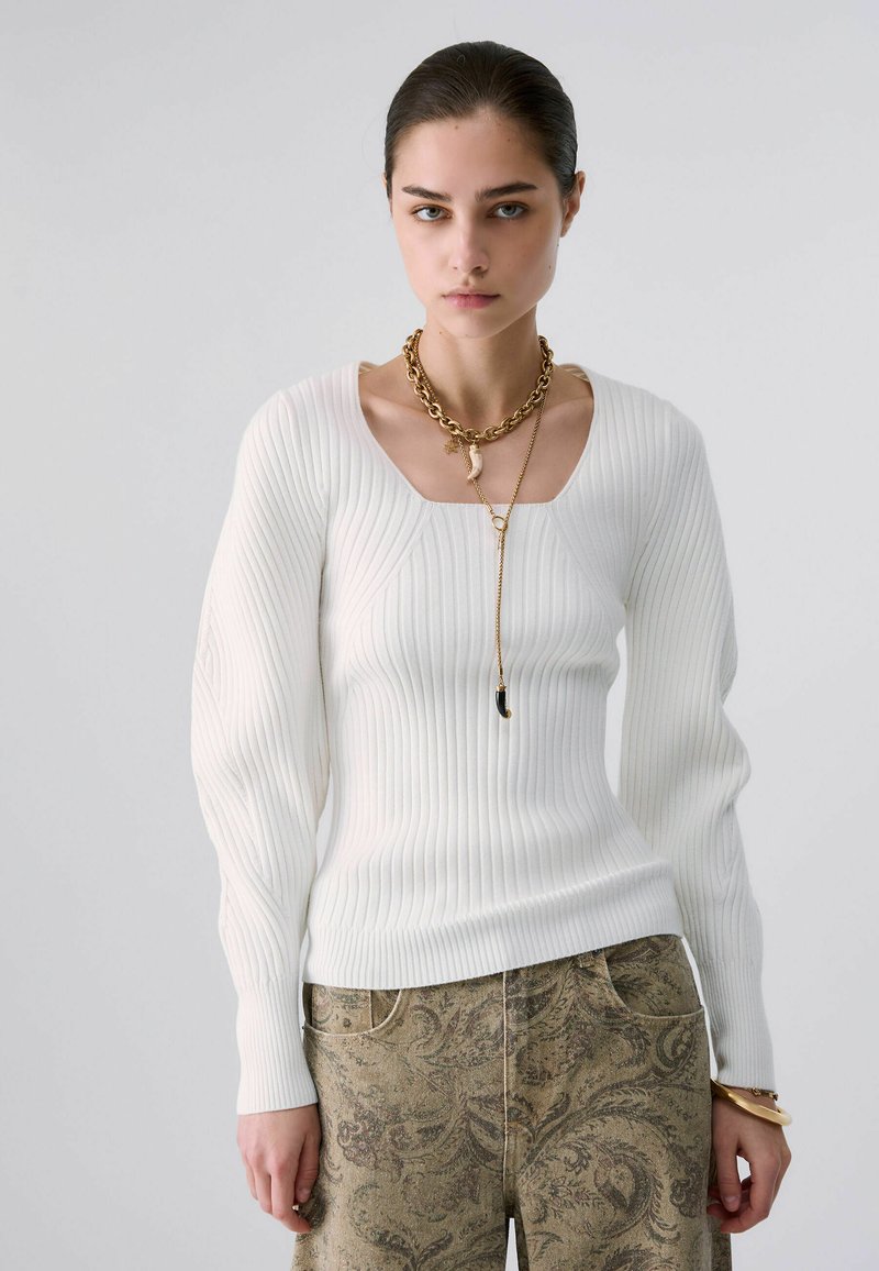 Jeune femme aux cheveux tirés en arrière, portant un pull blanc à côtes, un pantalon à motifs et des colliers en or superposés sur un fond gris uni.