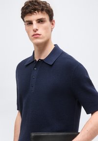 Νεαρός άνδρας με κοντά καστανά μαλλιά φοράει μπλε ναυτικού χρώματος πόλο μπλουζάκι με υφή και κρατάει μια μαύρη δερμάτινη τσάντα χειρός σε απλό ανοιχτόχρωμο φόντο.