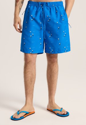 Niebieskie kąpielówki o lekkiej, gładkiej teksturze; wzorowane na małych surferach i falach. W połączeniu z niebieskimi i pomarańczowymi klapkami.