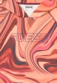 Tkanina w różowo-brzoskwiniowy abstrakcyjny wzór wiru z napisem "GUESS" wydrukowanym dużymi, półprzezroczystymi literami w pobliżu dekoltu.