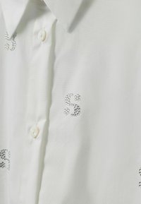 Hvid skjorte med knapper, der har sølv rhinsten-detaljer i et blomstermønster. Stoffet har en glat tekstur og et klassisk kravedesign.