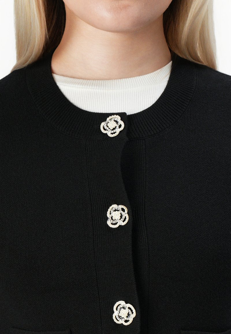 Cardigan noir avec trois boutons décoratifs en perles et rhinestones en forme de fleur sur un top à rayures blanc, porté par une personne à la peau claire.