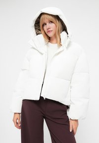 Jachetă puf albă cu glugă, având închidere cu fermoar, design căptușit și un căptușel interior de culoare maro contrastant. Croială texturată, scurtă.