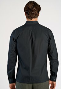 Zwarte longsleeve shirt van gladde stof, met een standaardkraag en knopenmanchetten. De achterkant heeft een subtiele yoke-naaddetail.