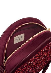 Borsa rotonda color bordeaux con una morbida texture in suede, dotata di chiusura con zip dorata, tasca interna e ricamo floreale sulla parte anteriore.