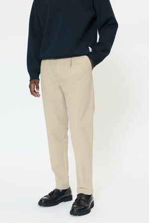 Pantalones beige plisados de tela, combinados con un suéter oscuro y zapatos negros que presentan un detalle de flecos texturizados.