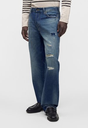 Homme portant un jean bleu déchiré à jambes larges, associé à des chaussures en cuir noir et une chemise rayée beige et noire à manches longues.
