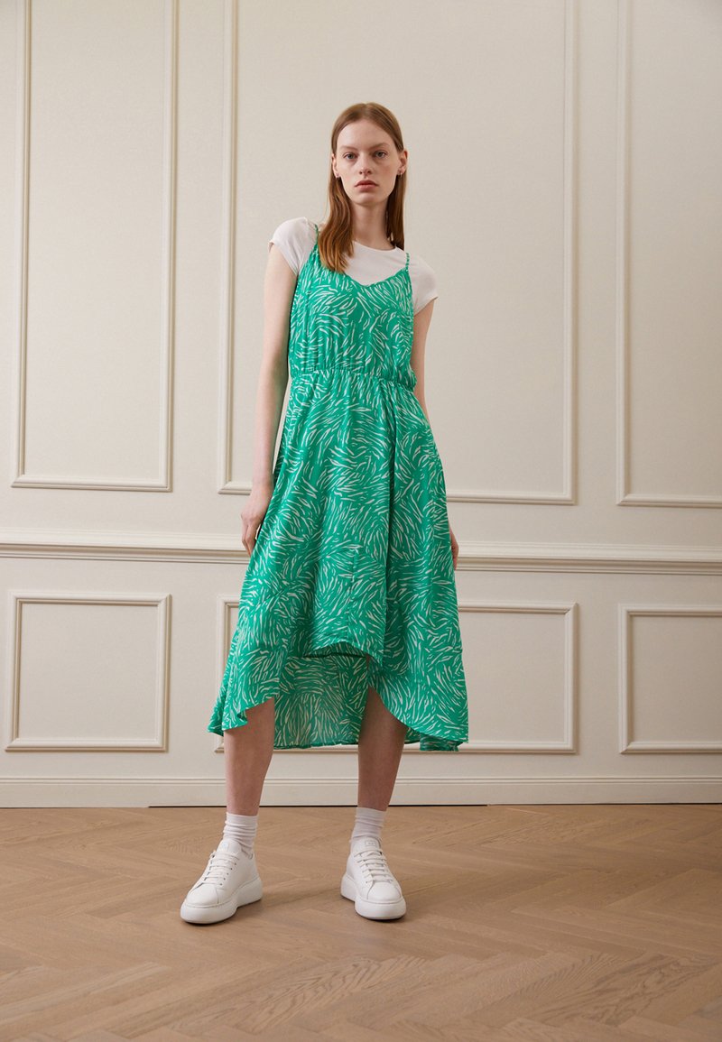 Vestido verde com padrão e design fluido, apresentando uma barra assimétrica. Combinado com ténis brancos e uma t-shirt branca justa por baixo.