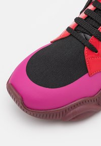 Baskets présentant une semelle en caoutchouc lisse rose, une tige en tissu noir texturé et des accents synthétiques rouges avec des lacets noirs.