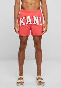 Coral swim shorts with white "KANI" lettering. Elastic waistband, side pockets, and a smooth texture. Worn with light-colored sandals.