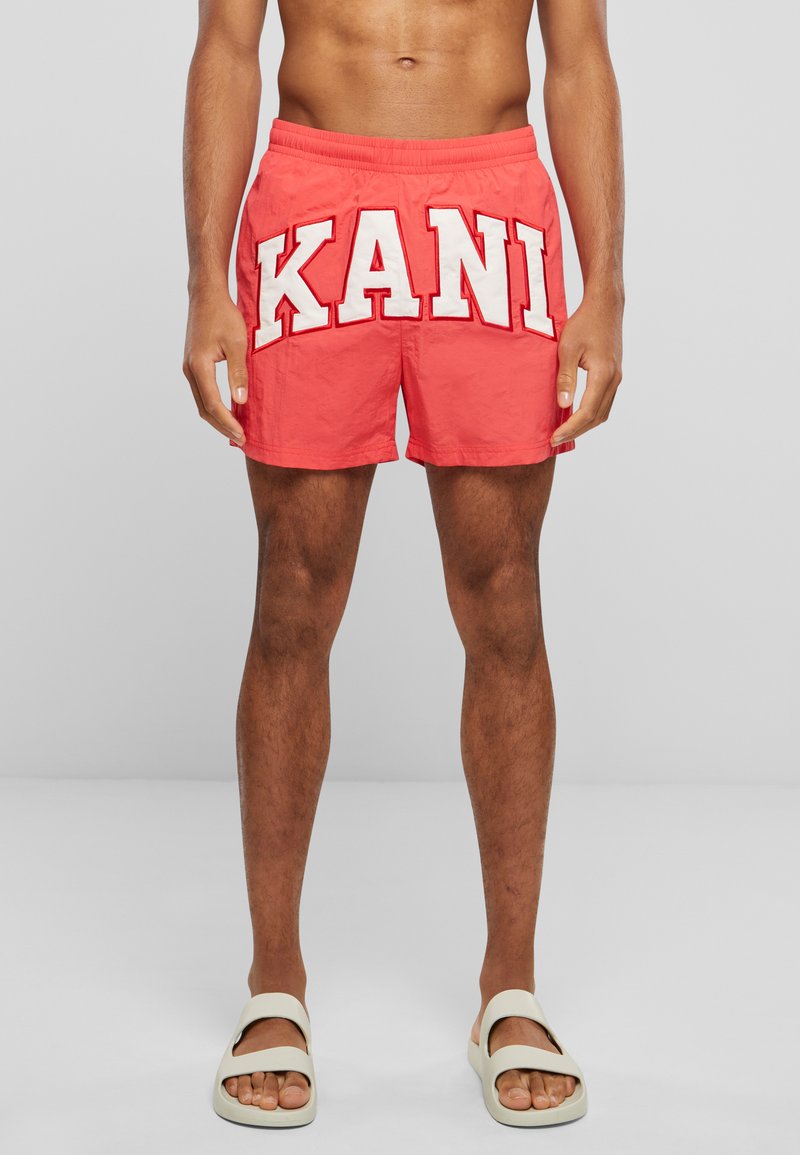 Coral swim shorts with white "KANI" lettering. Elastic waistband, side pockets, and a smooth texture. Worn with light-colored sandals.