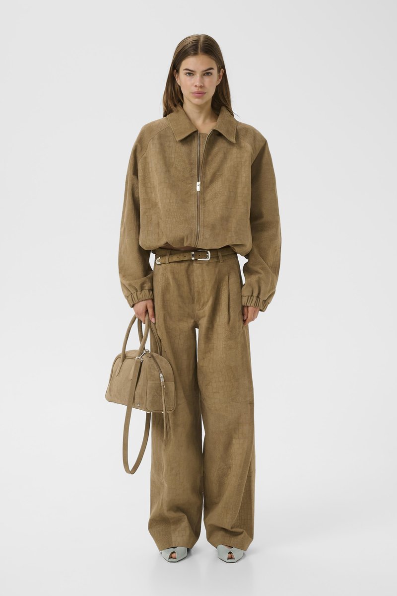 Nő barna texturált, egymáshoz illő cipzáras dzsekiben és bőszárú nadrágban, barna kézitáskát tart, fehér nyitott orrú cipőt visel.