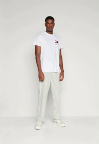 Άνδρας που στέκεται μπροστά σε απλό φόντο, φορώντας άσπρη μπλούζα με γραφικά, άσπρα παπούτσια και άσπρα παντελόνια με κατακόρυφες μαύρες ρίγες.