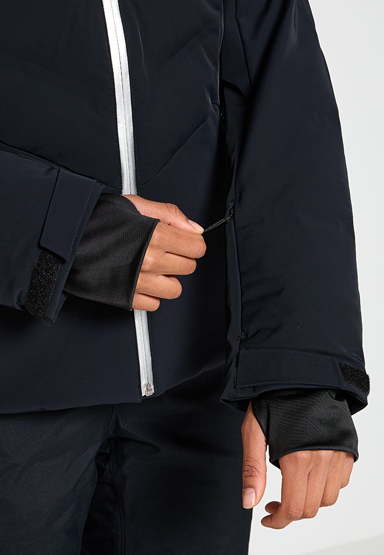 Zwarte geïsoleerde jas met witte rits; heeft klittenband manchetten en een verstelbare opening voor de pols voor een veilige pasvorm. Zachte textuur binnenvoering.