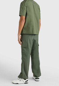 Persona con una camisa de manga corta verde oliva y pantalones cargo, con zapatillas blancas y negras, de pie contra un fondo claro y liso.