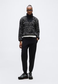 Persoană purtând un pulover cu model negru și bej, pantaloni sport negri, adidași gri și negri și ochelari de soare închiși la culoare, pe un fundal simplu.