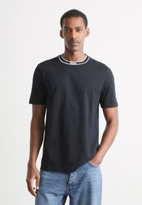 Juodas marškinėliai trumpomis rankovėmis, kontrastiniu baltu apykaklės kraštu ir logotipo detale ant apykaklės; derinami su mėlynais džinsais.