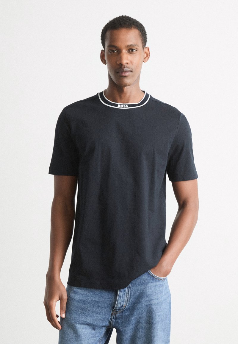 Juodas marškinėliai trumpomis rankovėmis, kontrastiniu baltu apykaklės kraštu ir logotipo detale ant apykaklės; derinami su mėlynais džinsais.