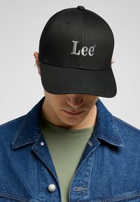 Black baseball cap with a curved brim, featuring grey embroidered "Lee" logo. Paired with a blue denim jacket and olive green shirt.