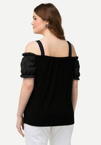 Top nero con design off-shoulder, maniche a palloncino in tessuto nero texturizzato e dettagli in pizzo sulle spalline. Design posteriore semplice, indossato con pantaloni bianchi.