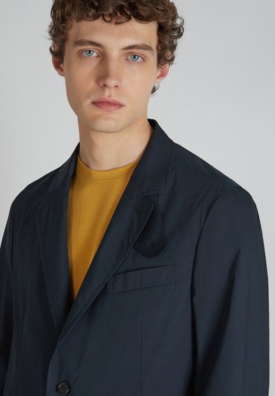 Blazer blu scuro realizzato in tessuto liscio con rever a punta, chiusura a un bottone e una piccola tasca sul petto. Indossato sopra una camicia senape.