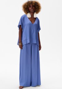 Niebieski satynowy komplet składający się z luźnej bluzki z dekoltem w serek i szerokich spodni. Posiada krótkie rękawy oraz swobodny krój zapewniający wygodę.