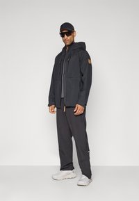 Mann iført sort hættejakke, sorte bukser, sort kasket, store solbriller og hvide sneakers, der står imod en ensfarvet grå baggrund.