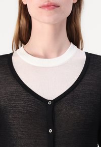 Svart stickad cardigan med V-ringning och knappstängning, över en vit ribbad kragtop. Mjuk textur, lätt design.