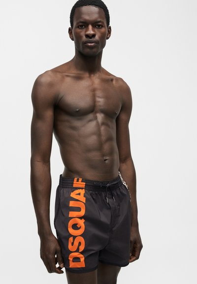 Άνδρας χωρίς πουκάμισο με καλογυμνασμένους μυς που φοράει μαύρα μαγιό με μεγάλο κάθετο πορτοκαλί κείμενο "DSQUARED" στην αριστερή πλευρά σε απλό φόντο.