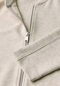 Detailní záběr na světle šedou mikinu na zip, která ukazuje zip a detaily rukávů, zdůrazňující měkkou texturu a prošívání.