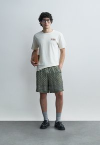 Mees lokkis juuste ja prillidega, valges t-särgis, mustriga rohelised lühikesed püksid, hallid sokid, mustad kingad, hoiab käes väikest pruuni kotti.