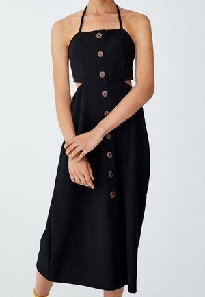 Ärmelloses schwarzes Midikleid mit Neckholder, vorderen Holzknöpfen und seitlichen Ausschnitten an der Taille, getragen von einer Person mit verschränkten Händen.