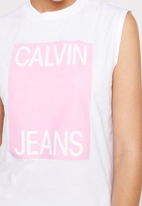 Bílý bezrukávový bavlněný tričko s růžovým obdélníkovým grafickým potiskem, na kterém je text "CALVIN JEANS" tučnými bílými písmeny.